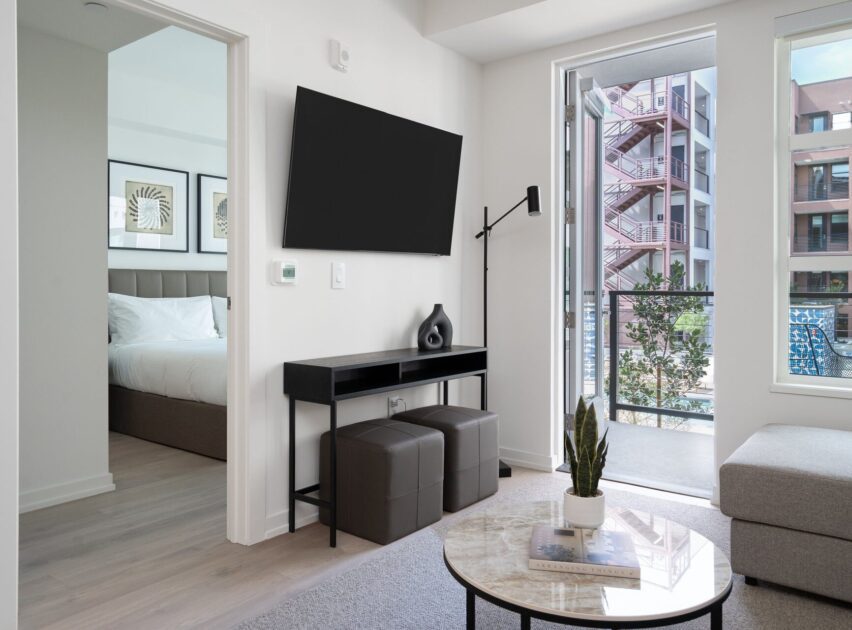 Modern apartment living room with a wall-mounted TV, console table, two ottomans, and a round coffee table with a plant. Bedroom with a bed is visible through an open door; balcony overlooks other buildings.
