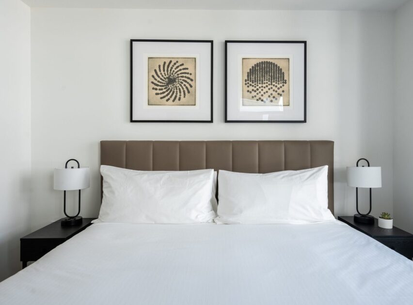 A modern bedroom with a neatly made bed, white bedding, a padded brown headboard, two black nightstands with white lamps, a small potted plant, and two framed abstract artworks above the headboard.