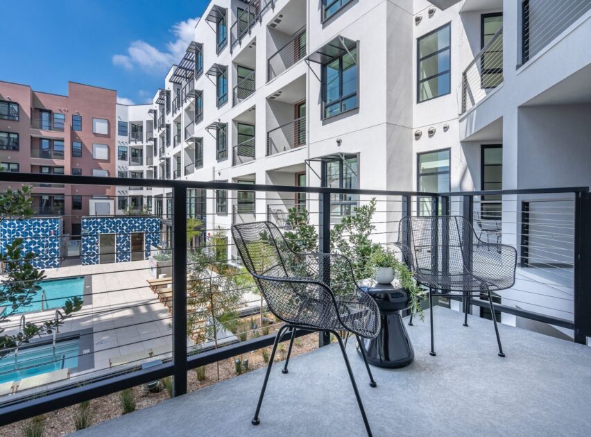 Modern apartment balcony with two black mesh chairs and a small table, overlooking a courtyard with a swimming pool and surrounding contemporary buildings under a blue sky.