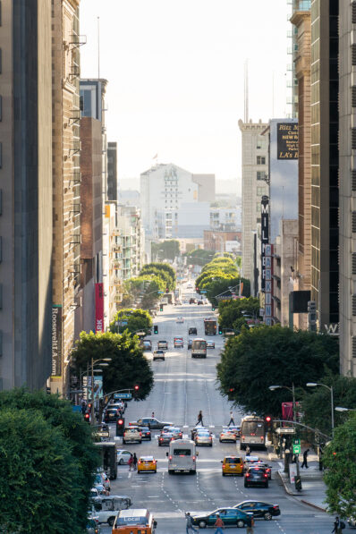 A busy city street in downtown Los Angeles with cars, taxis, and buses at an intersection, tall buildings on both sides, and trees lining the sidewalks under bright daylight.