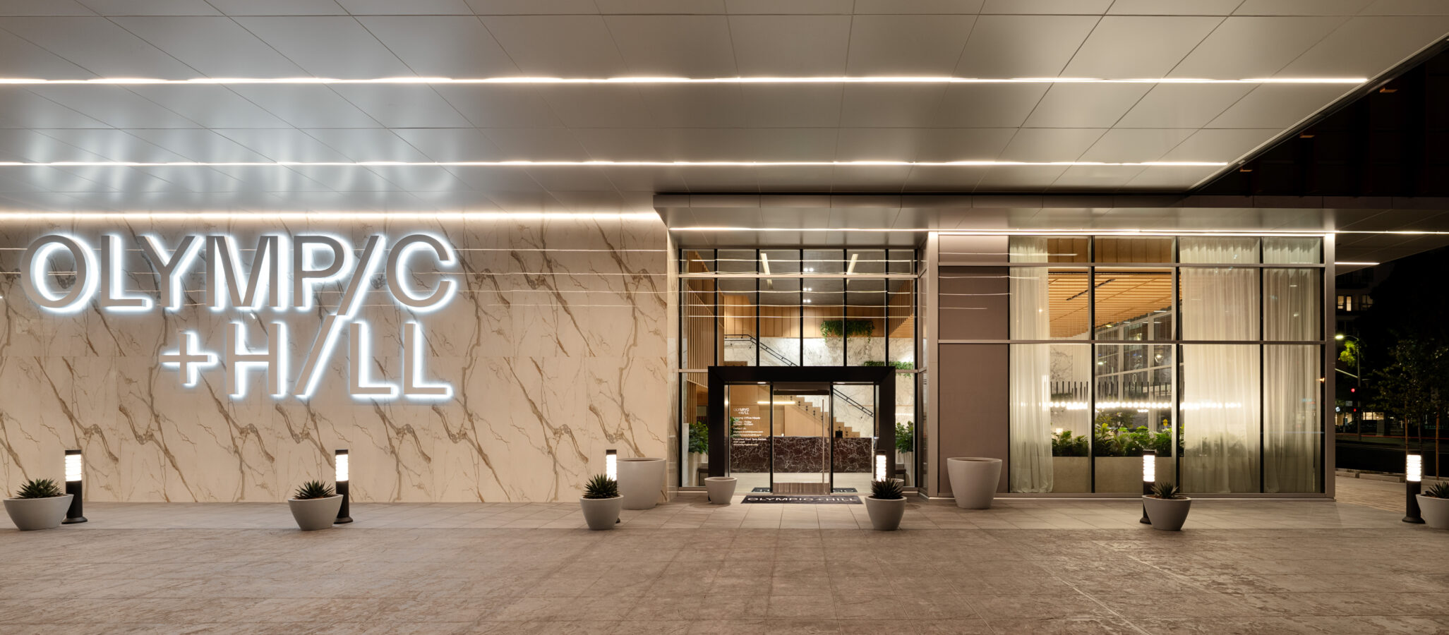 Exterior of a modern building with large illuminated letters spelling “OLYMPIC + HILL” on a marble wall, glass entrance doors, potted plants, and sleek overhead lighting.