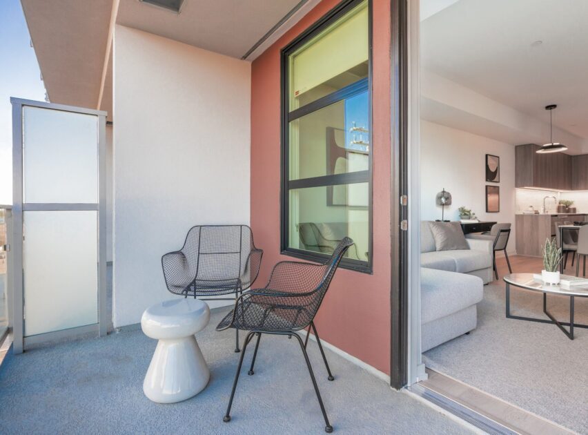 A modern apartment balcony with two black wire chairs and a small white table. The balcony overlooks an open living room with a gray sofa, a coffee table, and a kitchen area in the background.