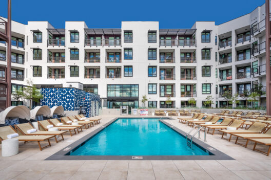 A modern apartment building with balconies surrounds a rectangular outdoor swimming pool. Lounge chairs and cabanas line the poolside, and the sky above is clear and blue.