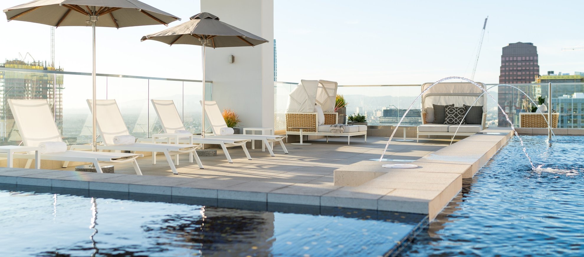 Rooftop pool area with lounge chairs, umbrellas, and modern seating beside clear blue water. City buildings and a crane are visible in the background under a bright sky.