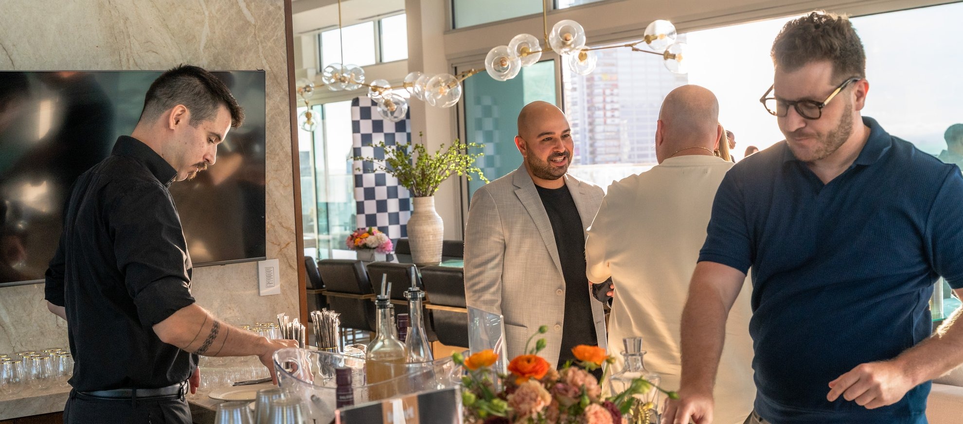 Four men are gathered in a modern, sunlit room with large windows. One man prepares drinks at a counter, while the others chat nearby. A vase of orange flowers and bottles sit on the counter in the foreground.
