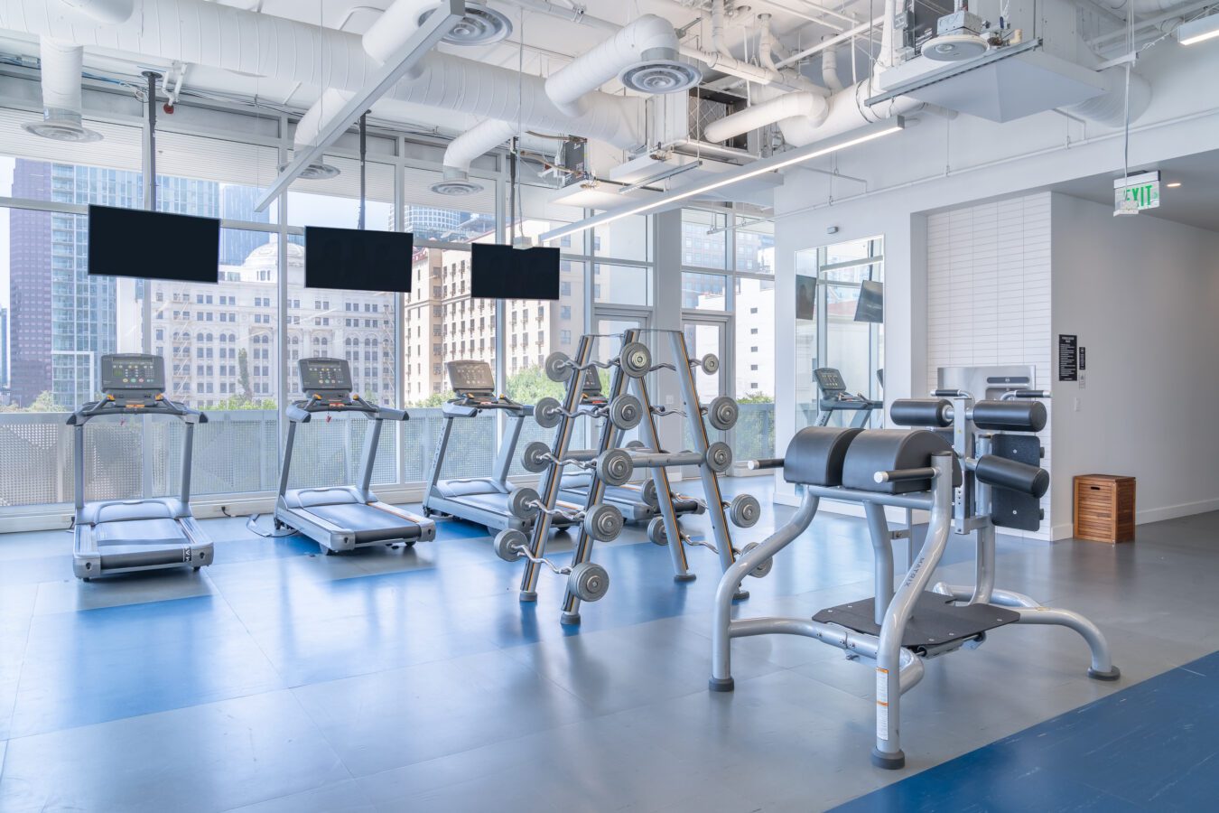 Modern gym with large windows, several treadmills, flat-screen TVs, dumbbell rack, and exercise benches; city buildings visible outside. The space is bright with blue and gray flooring.