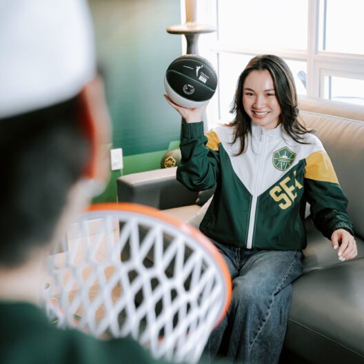 A woman in a green and white SEATTLE basketball jacket sits on a couch, smiling while holding a small basketball, aiming toward a person holding a toy basketball hoop in the foreground.
