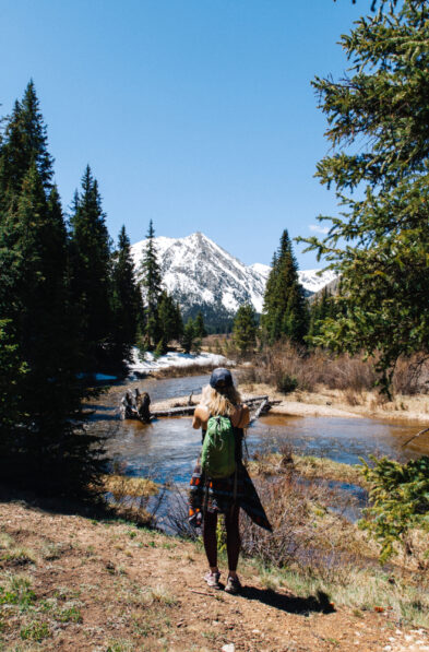 A person with a green backpack stands on a trail near a creek, facing snow-capped mountains and pine trees under a clear blue sky.