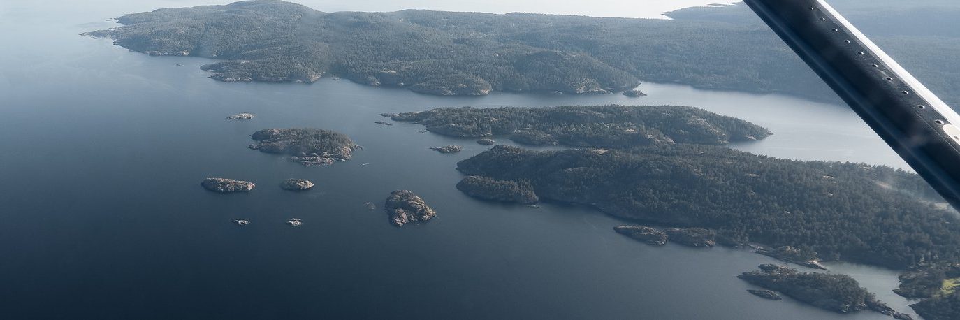 Aerial view of tree-covered islands in a large body of water, with a portion of an aircraft wing visible in the upper right corner. The scene appears calm and expansive.