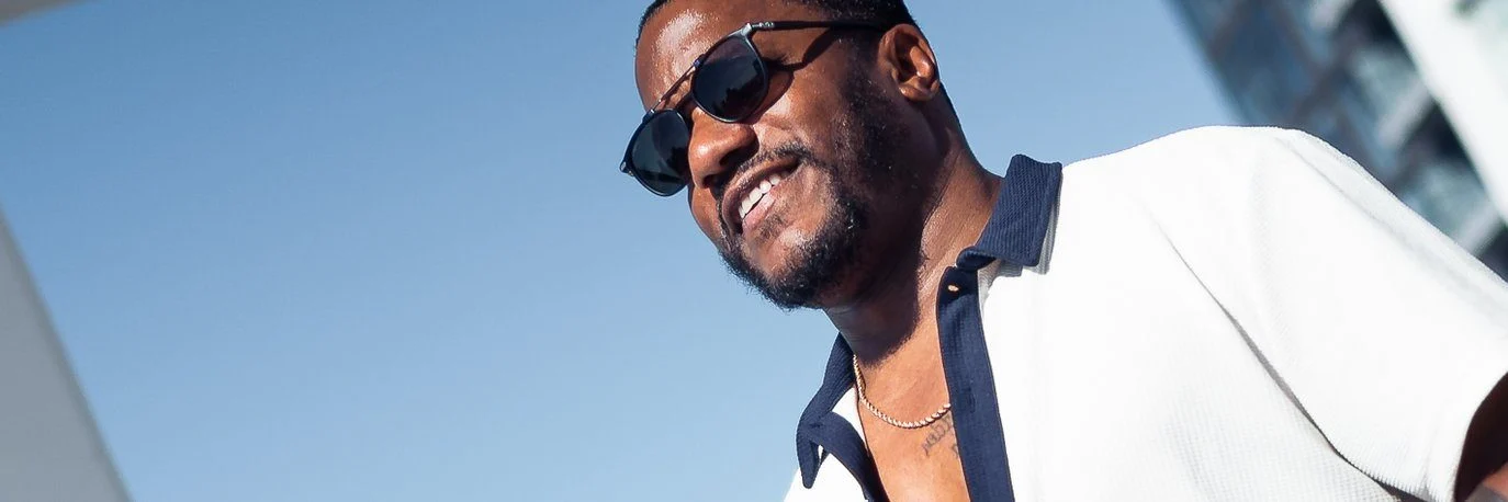 A man wearing sunglasses and a white shirt with a dark collar smiles outdoors on a sunny day, with a blue sky and a modern building in the background.