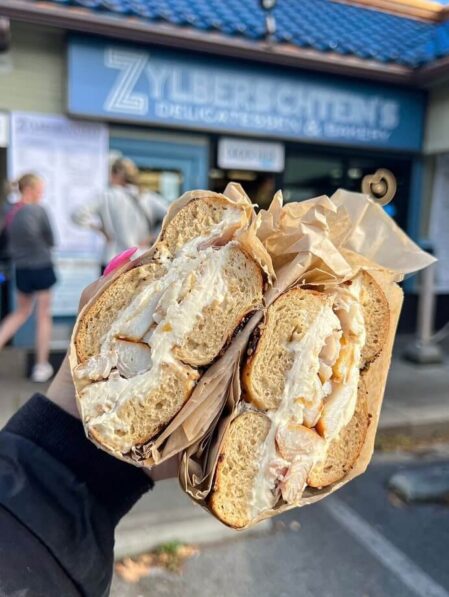 A hand holds two halves of a bagel sandwich filled with cream cheese and what appears to be smoked fish, with a deli storefront and people in the background.