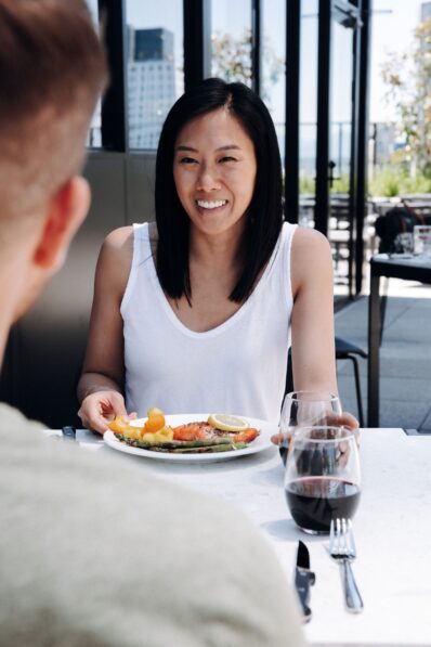 A woman in a white tank top smiles while dining outdoors, holding a plate of food with vegetables and lemon. Two glasses of red wine are on the table, and another person is seated across from her.