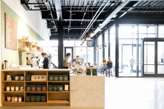 Bright modern coffee shop with large windows, a marble counter, shelves with coffee bags and mugs, baristas preparing drinks, and customers sitting at tables near the entrance.