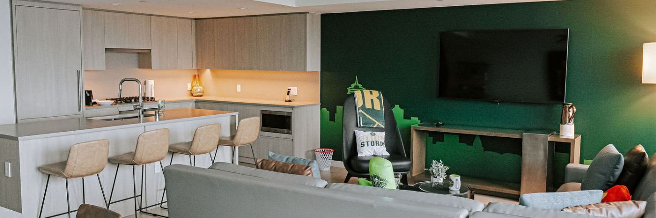 Modern open-plan living area with a kitchen featuring light wood cabinets, barstools, and a living room with a gray couch, wall-mounted TV, and green accent wall decorated with a cityscape mural.