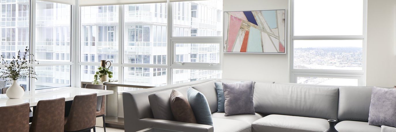 Modern living room with large windows, gray sectional sofa, and dining area with brown chairs. Abstract art hangs on the wall, and city buildings are visible outside through the bright windows.