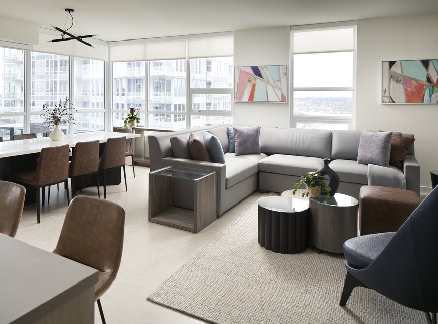 A modern, bright living and dining area with large windows, a gray sectional sofa, a dining table with brown chairs, abstract wall art, and a neutral color palette.