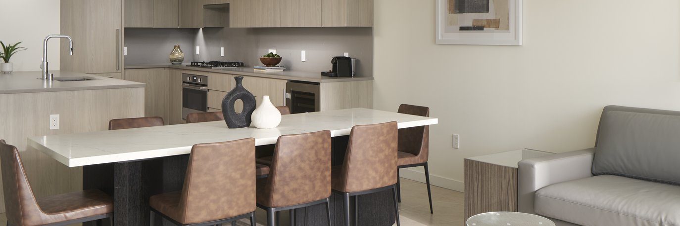 Modern kitchen and dining area with a marble dining table, six brown chairs, minimalist decor, light wood cabinets, built-in appliances, and an adjacent living space with a gray sofa and wall art.