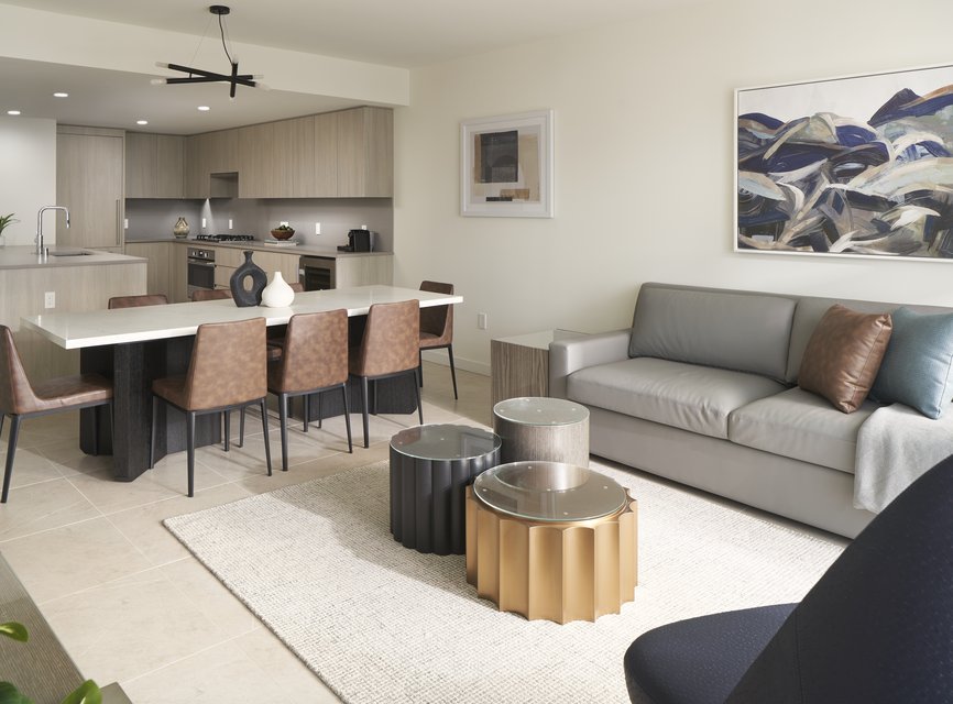 Modern open-plan living room and kitchen with neutral tones; features a gray sofa, abstract art, unique gold and black coffee tables, a dining table with brown chairs, and light wood cabinetry.