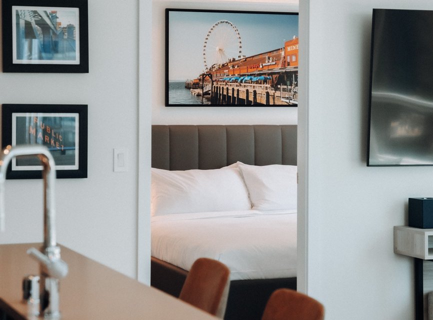 A modern hotel room with a neatly made bed seen through a doorway, wall art featuring a ferris wheel and pier, and part of a kitchen counter with a faucet in the foreground.