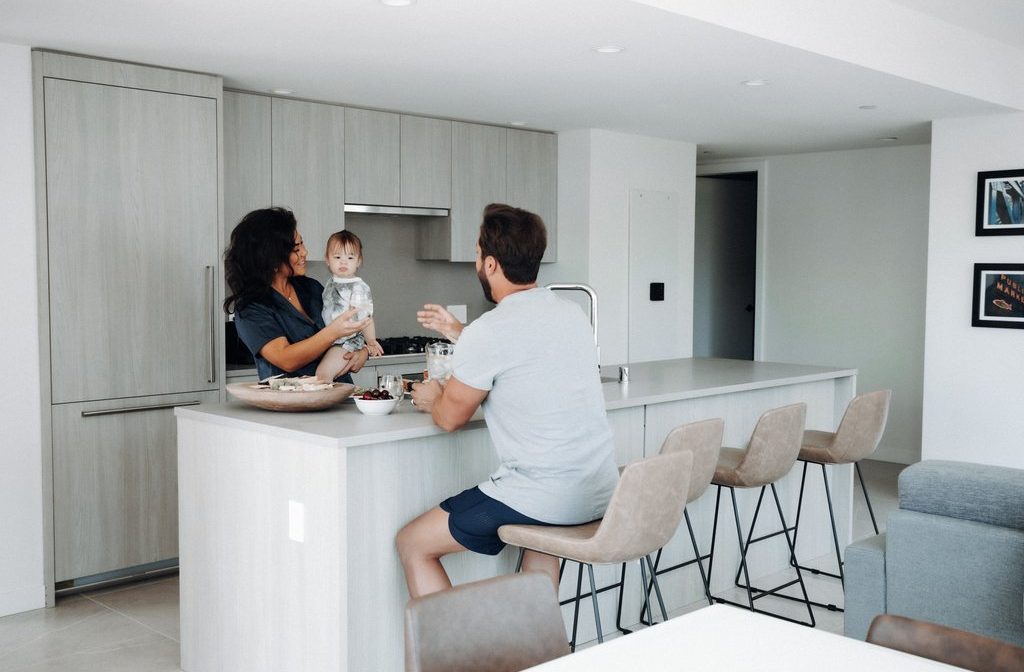 A woman holding a baby stands in a modern kitchen, talking to a man sitting on a barstool at the island counter. The kitchen is bright with light wood cabinets and neutral décor.