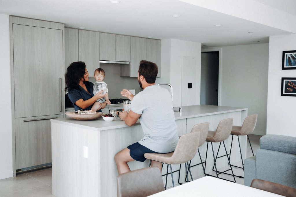 A woman holds a baby while talking to a man seated at a modern kitchen island. The kitchen features light wood cabinets, bar stools, and minimalistic decor.