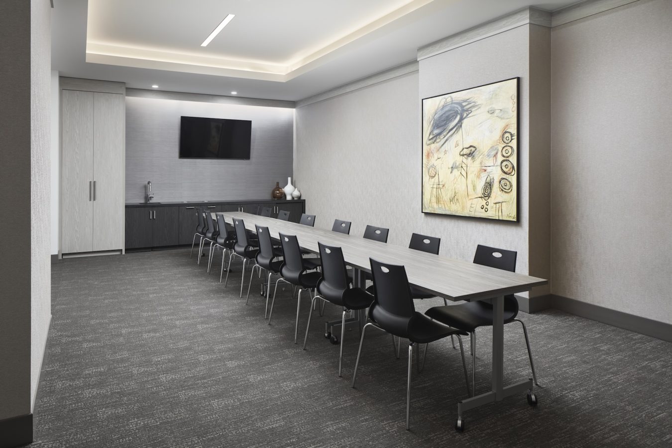 A modern, empty conference room with a long rectangular table, ten black chairs, a mounted flat-screen TV, abstract wall art, and cabinets along the back wall under soft ceiling lighting.