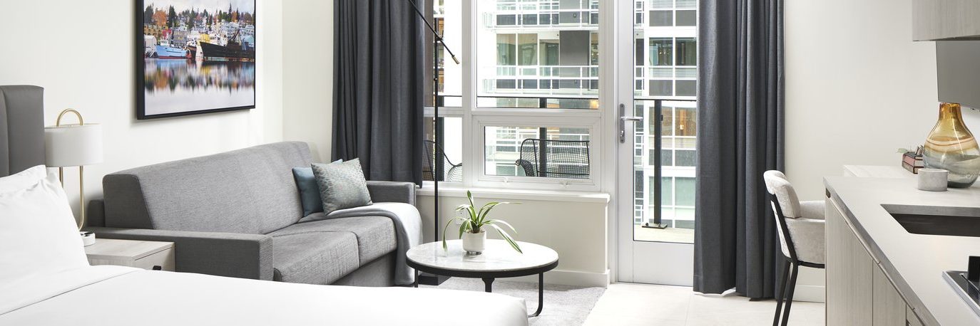 Modern studio apartment with a bed, gray sofa, round coffee table, and kitchenette. Large window with dark curtains opens to a balcony overlooking other contemporary apartment buildings.
