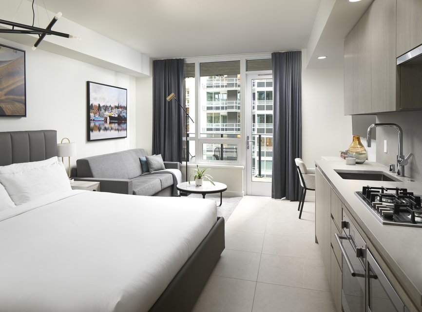 Modern studio apartment with a bed, gray sofa, round coffee table, wall art, kitchenette with stove and oven, and large windows with dark curtains overlooking a city building.