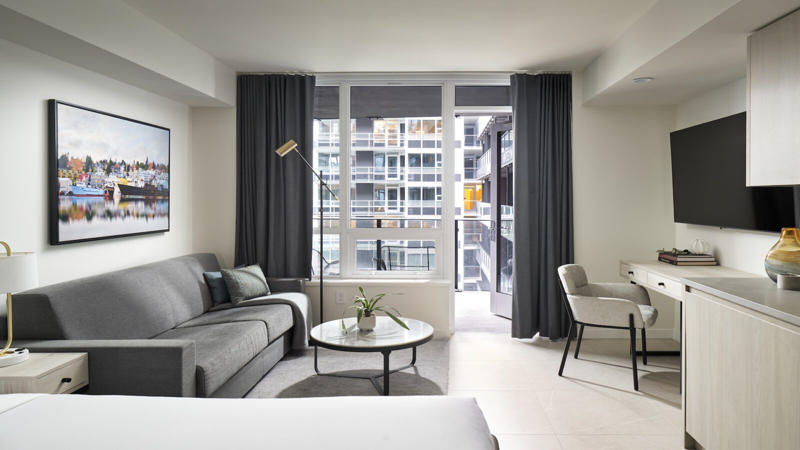 Modern hotel room with a gray sofa, round coffee table, desk with chair, flat-screen TV, and large window with blackout curtains, offering a view of neighboring buildings. A cityscape painting hangs above the sofa.