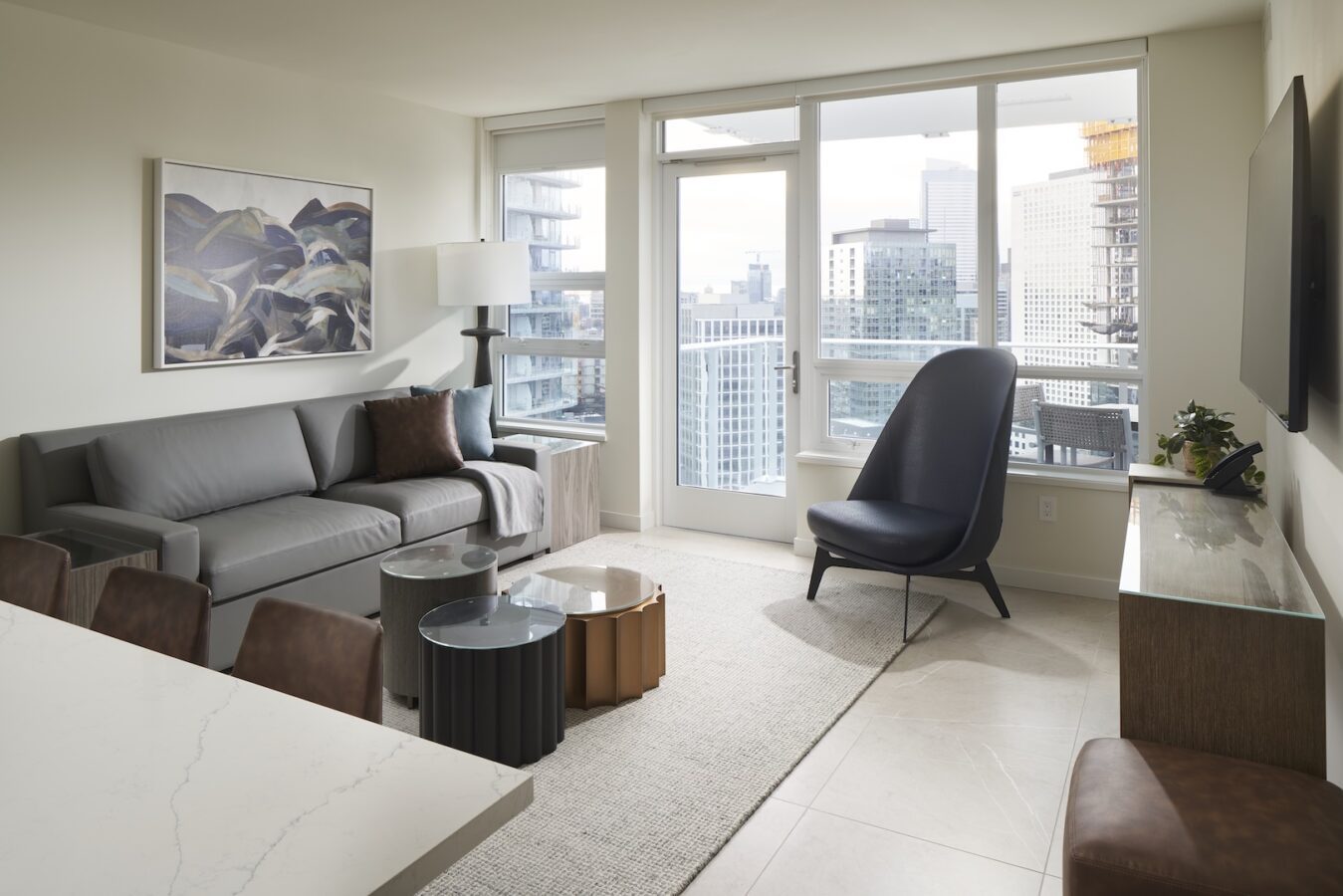 Modern living room with a gray sofa, black armchair, coffee tables, floor lamp, wall art, and large windows showing a cityscape with tall buildings. TV mounted on wall and neutral decor throughout.