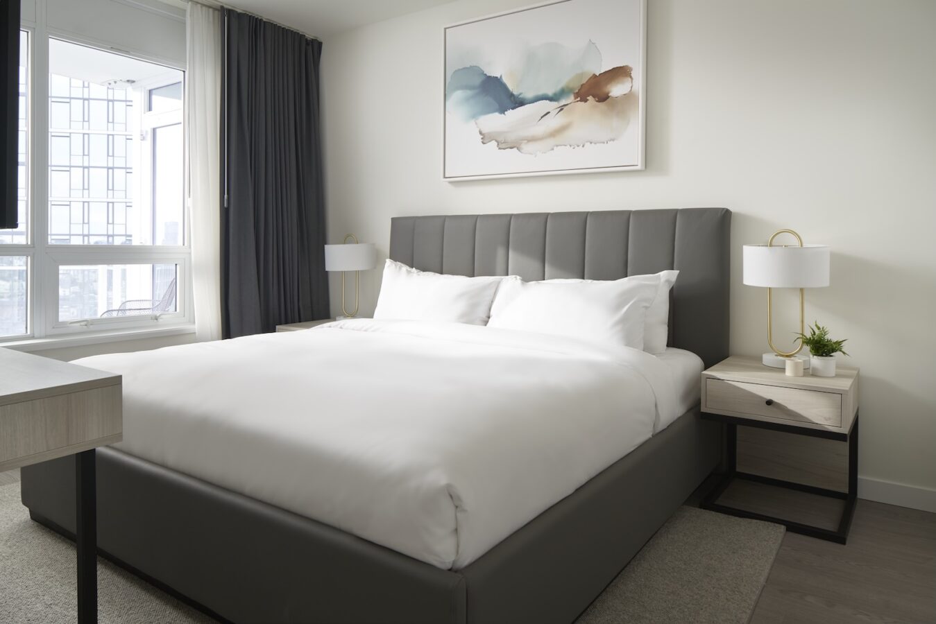 Modern bedroom with a large bed covered in white linens, two bedside tables with lamps and a small plant, a large window with dark curtains, and abstract art on the wall above the headboard.