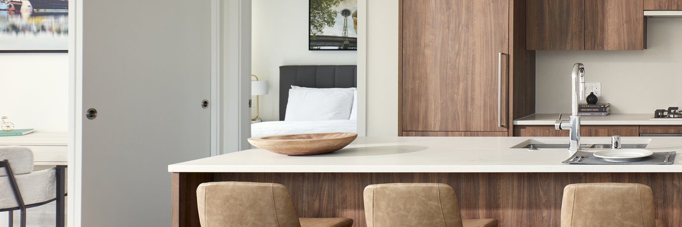 Modern kitchen with a white countertop, wooden cabinets, and three tan barstools. A wooden bowl sits on the counter. In the background, a bedroom with a white bed is visible through a doorway.