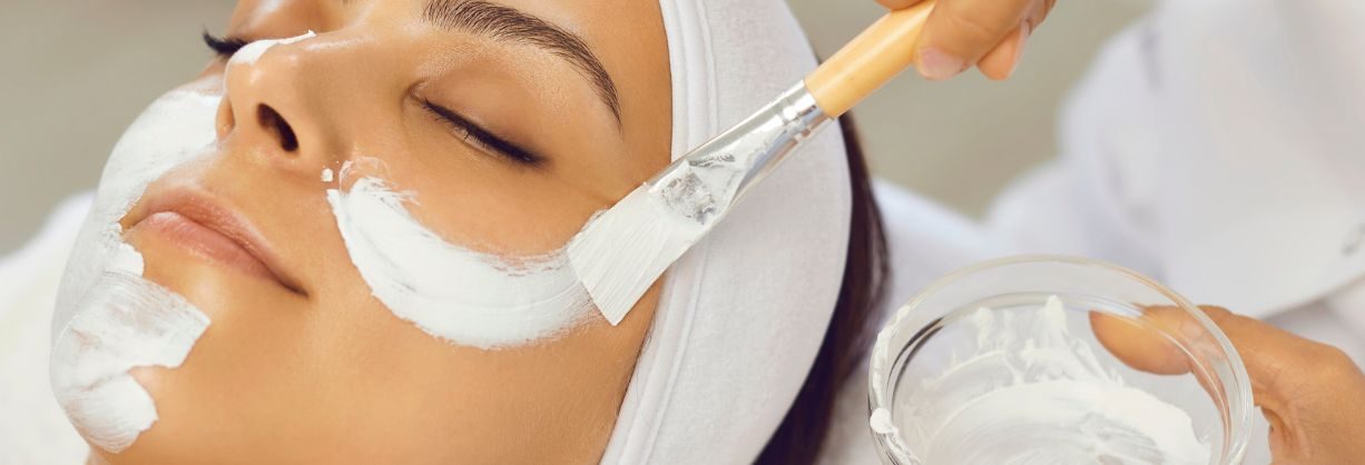 A person with closed eyes is lying down with a white headband while a brush is used to apply a white facial mask to their face. The setting appears to be a spa or skincare clinic.