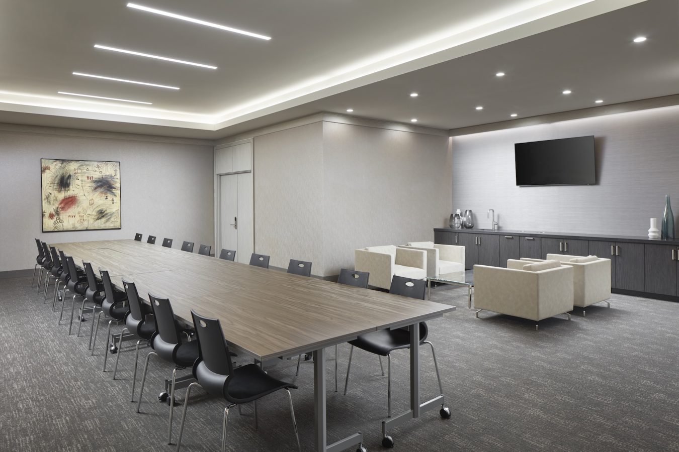 Modern conference room with a long rectangular table and black chairs, a lounge area with beige armchairs, a wall-mounted TV, cabinets, and a piece of abstract artwork on the wall. The room is well-lit with ceiling lights.
