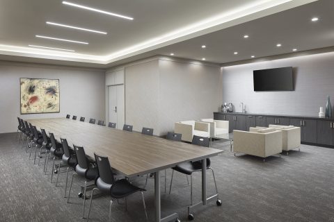 Modern conference room with a long rectangular table and black chairs, a lounge area with beige armchairs, a wall-mounted TV, cabinets, and a piece of abstract artwork on the wall. The room is well-lit with ceiling lights.