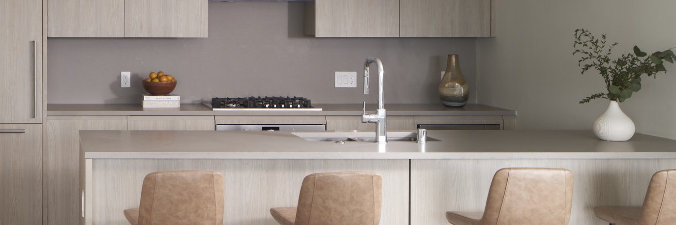Modern kitchen with light wood cabinets and island, beige bar stools, a built-in stove, a stainless steel sink, and simple decor including a bowl of fruit and a vase with greenery.