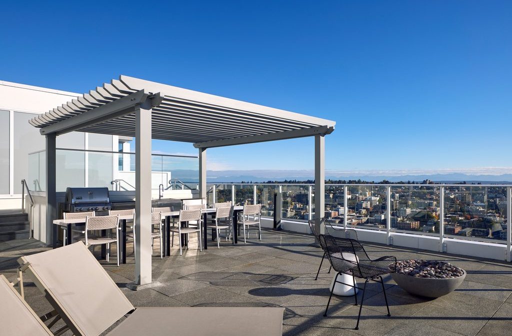 A modern rooftop patio with a pergola, outdoor dining table, grill, lounge chairs, and a fire pit, overlooking a cityscape under a clear blue sky. Glass railings surround the spacious, sunlit area.