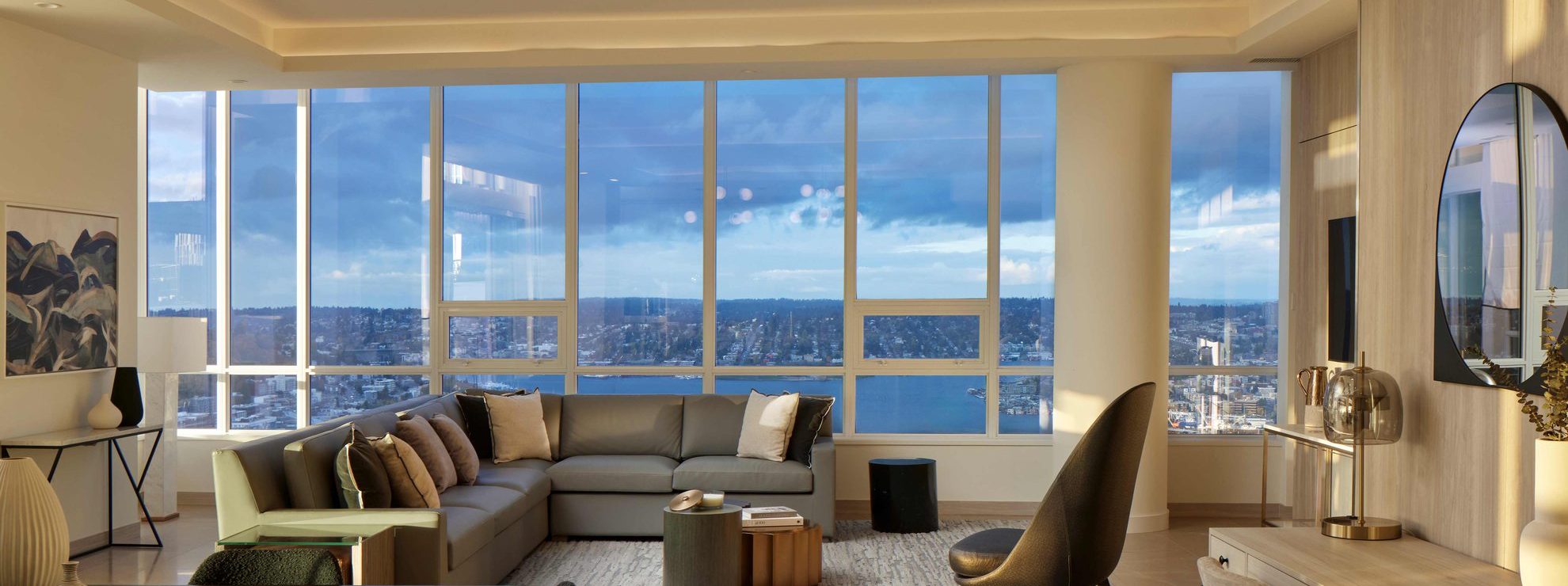Modern living room with floor-to-ceiling windows, offering a scenic view of a river and city. The space features a large gray sectional sofa, stylish decor, and neutral tones with natural light filling the room.