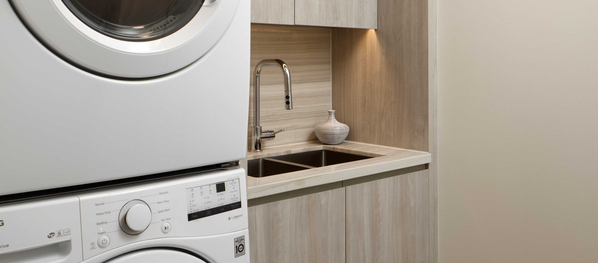 A modern laundry room with a stacked washer and dryer next to a built-in sink, a chrome faucet, light wood cabinets, and a small decorative vase on the countertop.