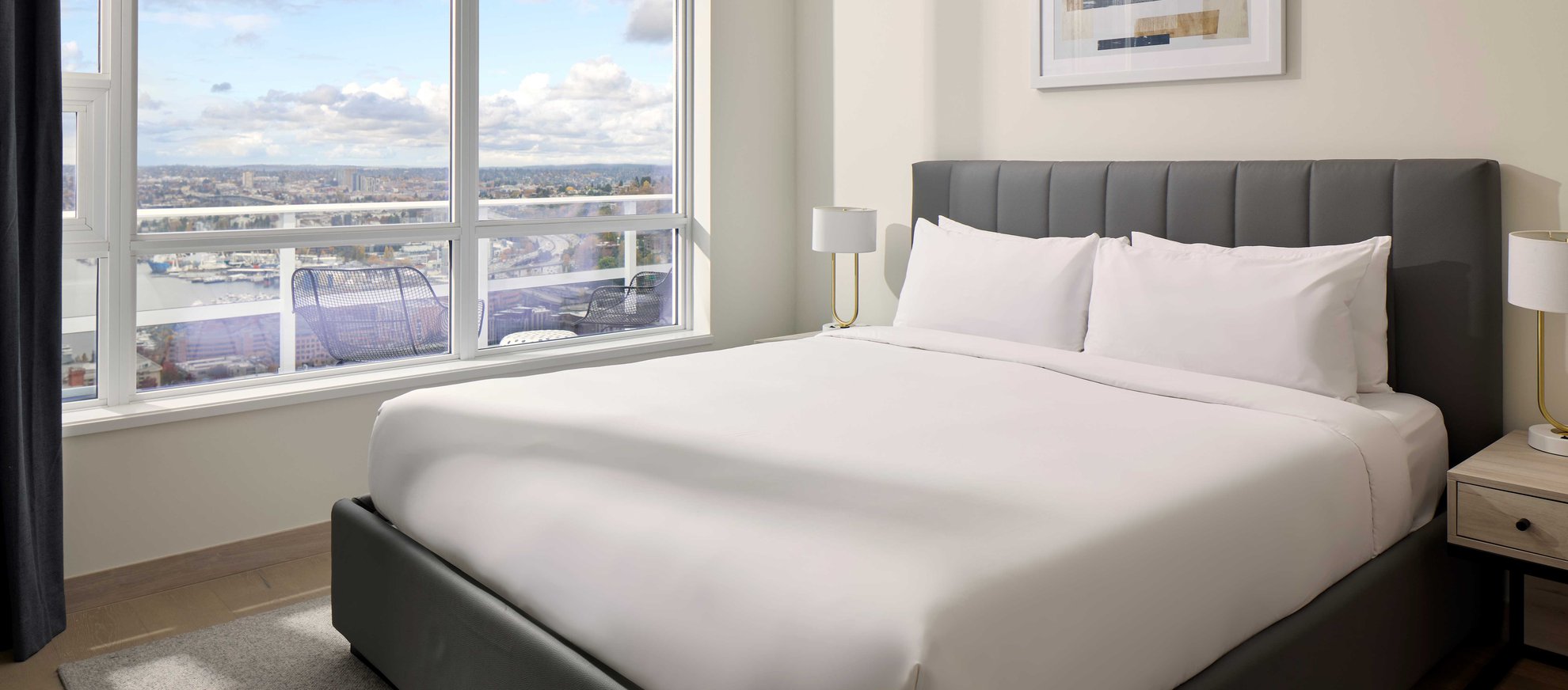 A modern bedroom with a neatly made bed, two bedside tables with lamps, and a large window offering a city and water view on a bright, partly cloudy day.