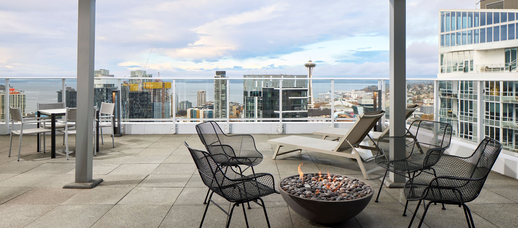 A modern rooftop patio with black wire chairs around a fire pit, lounge chairs, and a table, overlooking a city skyline, water, and distant buildings under a cloudy sky.