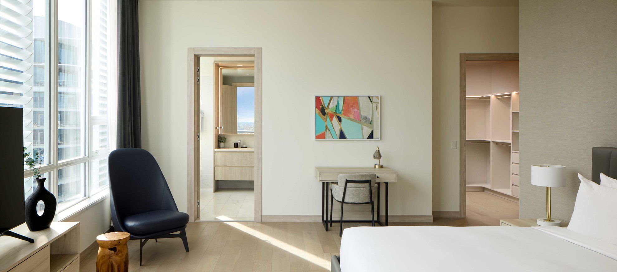 A modern, bright bedroom with a bed, a small desk and chair, an abstract painting, a dark armchair, large windows, and doors leading to a bathroom and a walk-in closet.