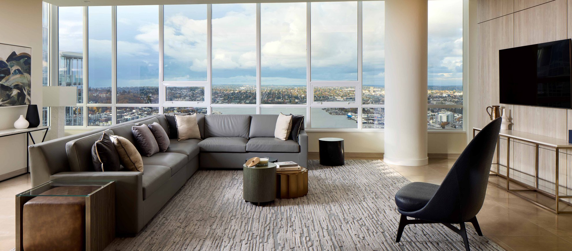 Modern living room with large floor-to-ceiling windows offering a city and water view, gray sectional sofa with pillows, armchair, rug, side tables, and wall-mounted TV, decorated in neutral tones.