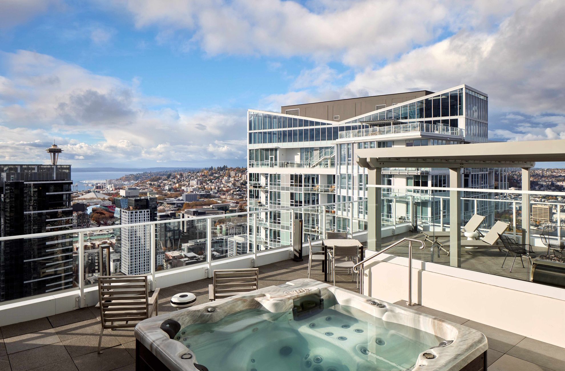 A rooftop patio with a hot tub, lounge chairs, and a glass railing overlooks a city skyline with modern buildings and distant water under a partly cloudy sky.