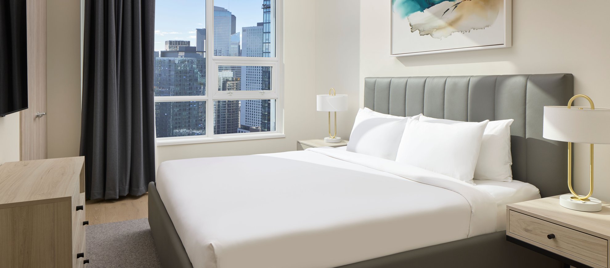 A modern bedroom with a large bed, gray headboard, white bedding, two bedside tables with lamps, a dresser, gray curtains, and a window overlooking city skyscrapers. Abstract art hangs above the bed.