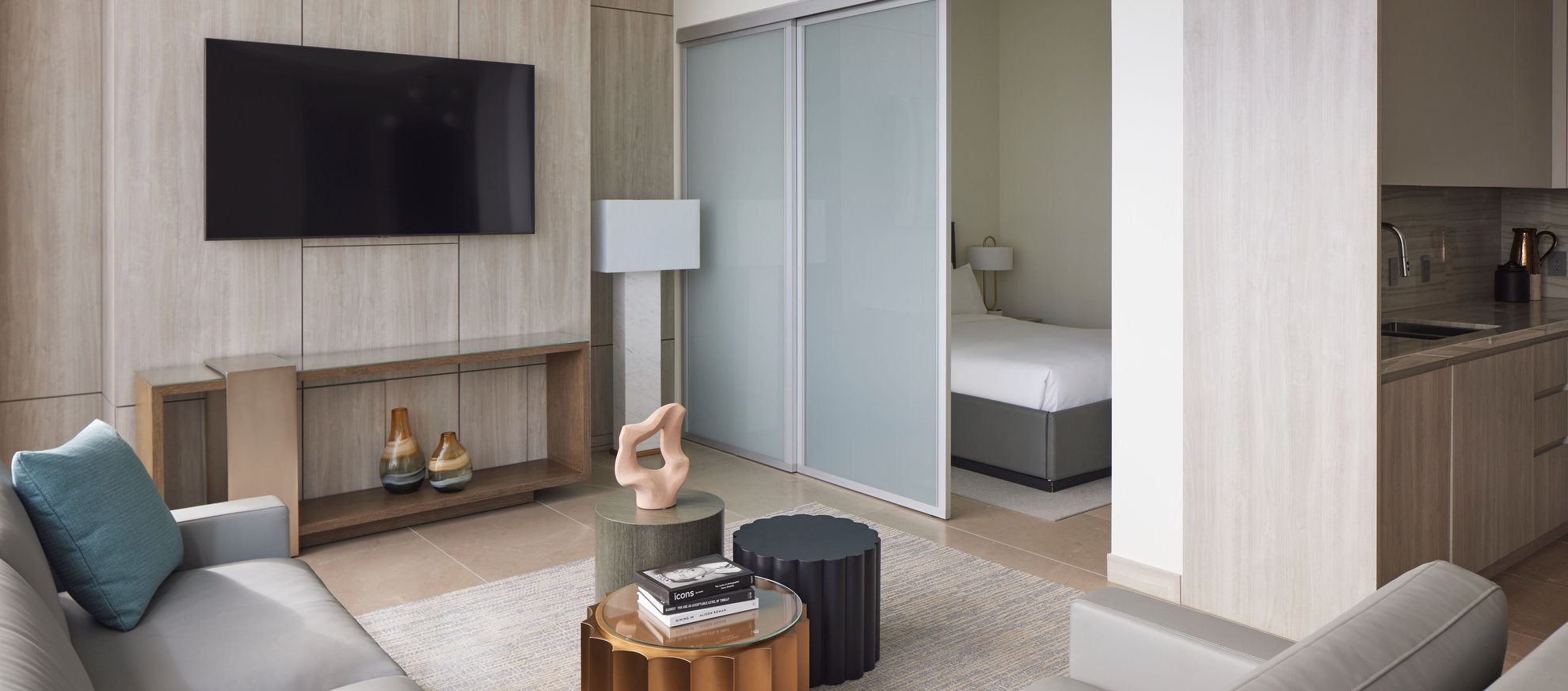 Modern living room with light wood decor, a wall-mounted TV, gray sofa, coffee tables with books and sculptures, and a view into a bedroom through sliding frosted glass doors; small kitchen area on the right.