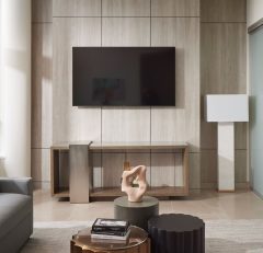 Modern living room with a grey sofa, large windows, wall-mounted TV, minimalist console table, decorative sculpture on a small round table, and a sliding door leading to a bedroom.