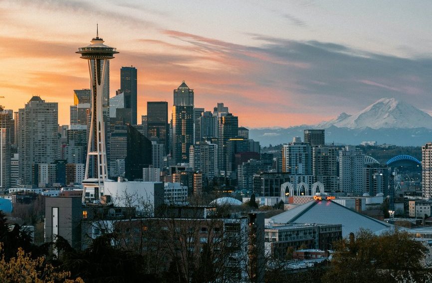 Seattle skyline at sunset, featuring the Space Needle, modern skyscrapers, and Mount Rainier in the background under a colorful sky with scattered clouds.