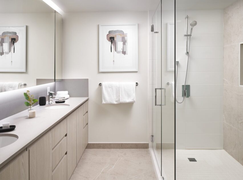 Modern bathroom with light wood cabinets, double sinks, a glass-enclosed shower, white towels on a rack, taupe tile floor, and abstract art on the wall above the towel rack.