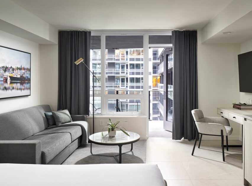 Modern hotel room with a gray sofa, round coffee table, floor lamp, and large window with dark curtains overlooking city buildings. Desk with chair and TV are on the right. Neutral tones and minimal decor.